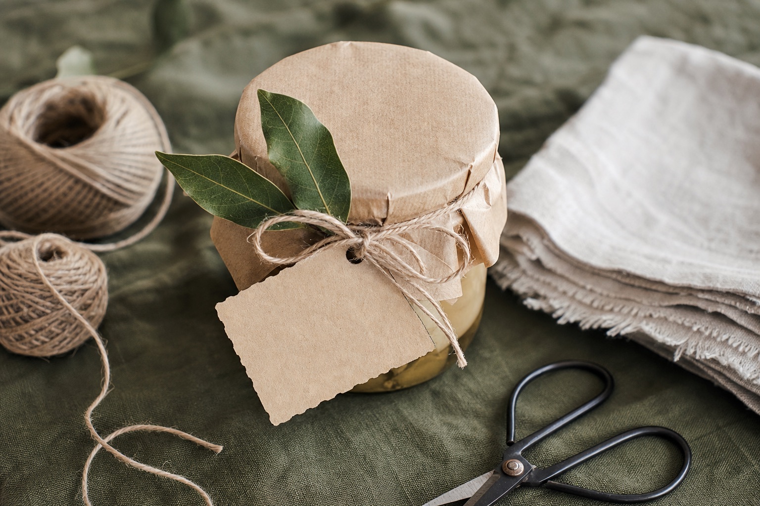 A kraft-wrapped glass jar tied with twine, with a dried bay sprig, a small blank card, a folded linen square and scissors